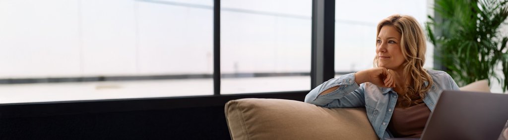 Une femme est assise sur un canap&eacute; avec un ordinateur portable et regarde par la fen&ecirc;tre | Eine Frau sitzt mit einem Laptop auf einem Sofa und schaut aus dem Fenster.