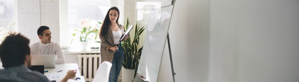 Eine junge Frau steht vor einem Whiteboard und pr&auml;sentiert Informationen. Zwei M&auml;nner sitzen an einem Tisch und h&ouml;ren ihr aufmerksam zu. | Une jeune femme se tient devant un tableau blanc et pr&eacute;sente des informations. Deux hommes sont assis &agrave; une table et l'&eacute;coutent attentivement. | A young woman stands in front of a whiteboard and presents information. Two men are sitting at a table and are listening to her attentively.