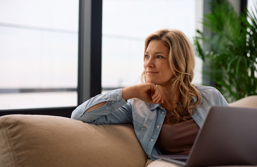 Une femme est assise sur un canapé avec un ordinateur portable et regarde par la fenêtre | Eine Frau sitzt mit einem Laptop auf einem Sofa und schaut aus dem Fenster.