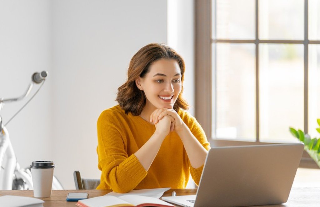 Eine junge Frau im orangen Pullover arbeitet an ihrem Laptop. | Une jeune femme en pull orange travaille sur son ordinateur portable. | A young woman in an orange sweater is working on her laptop.
